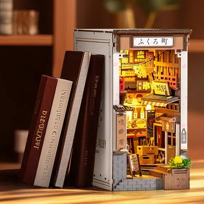 A completed Robotime Sakura Wine Alley Book Nook glowing with warm LED lights between books on a shelf, showing a detailed Japanese miniature street.