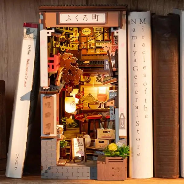 A completed Robotime Sakura Wine Alley Book Nook glowing with warm LED lights between books on a shelf, showing a detailed Japanese miniature street.