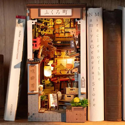 A completed Robotime Sakura Wine Alley Book Nook glowing with warm LED lights between books on a shelf, showing a detailed Japanese miniature street.